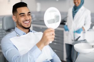 Man in dental chair smiling at perfect teeth in mirror with dentist blurry in background